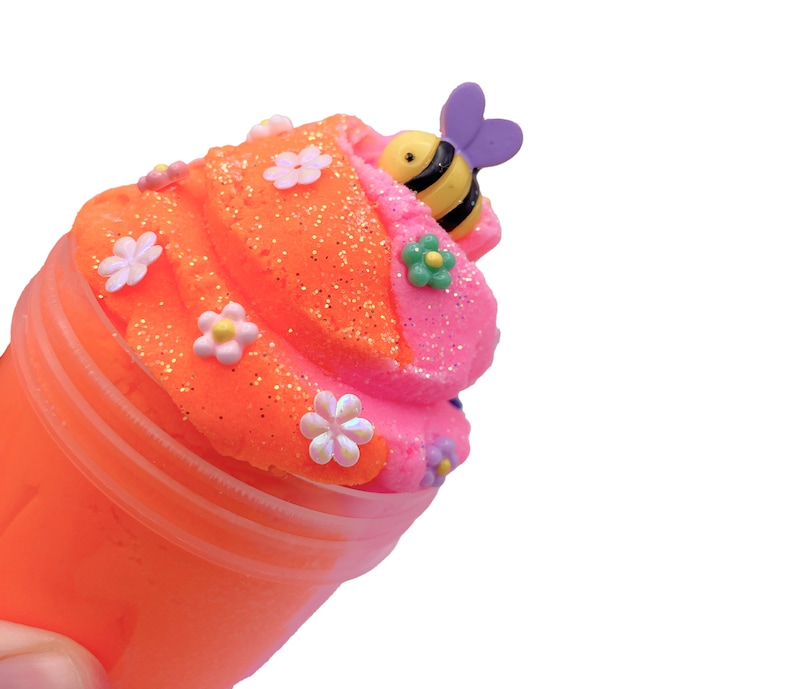 K&ouml;nnte beinhalten: Orangefarbener und rosafarbener Schleim in einem durchsichtigen Plastikbecher mit Bienen- und Blumenverzierungen.