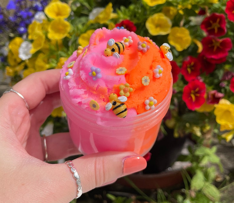 K&ouml;nnte beinhalten: Ein rosa und orangefarbener Schleim mit einem Wirbeldesign, verziert mit kleinen Blumen und Bienenanh&auml;ngern. Der Schleim befindet sich in einem durchsichtigen Plastikbeh&auml;lter.
