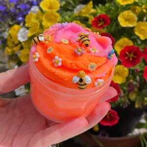 K&ouml;nnte beinhalten: Orangefarbener und rosafarbener Schleim mit Bienen- und Blumenverzierungen in einem durchsichtigen Plastikbeh&auml;lter.