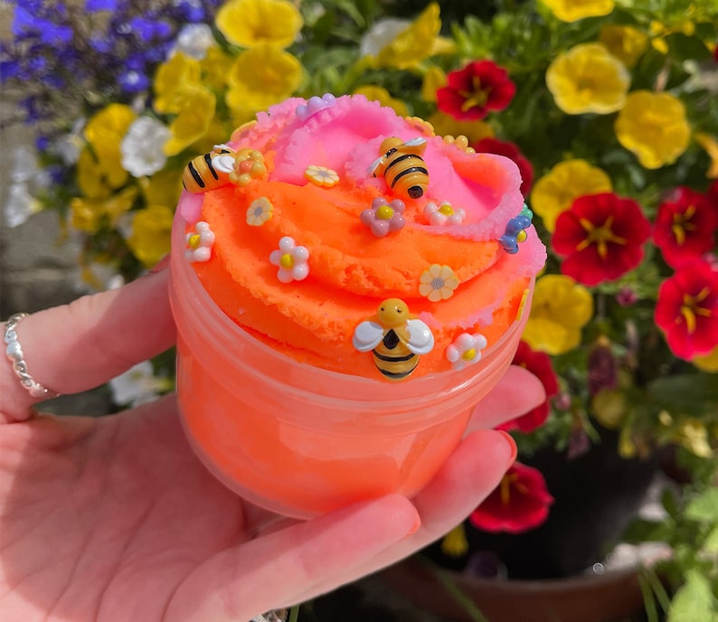 K&ouml;nnte beinhalten: Orangefarbener und rosafarbener Schleim mit Bienen- und Blumenverzierungen in einem durchsichtigen Plastikbeh&auml;lter.