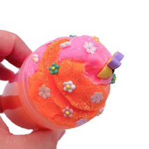 K&ouml;nnte beinhalten: Orangefarbener und rosafarbener Schleim mit Glitzer und wei&szlig;en Blumen in einem durchsichtigen Plastikbeh&auml;lter. Der Schleim ist mit einem gelben und violetten Blumenanh&auml;nger verziert.