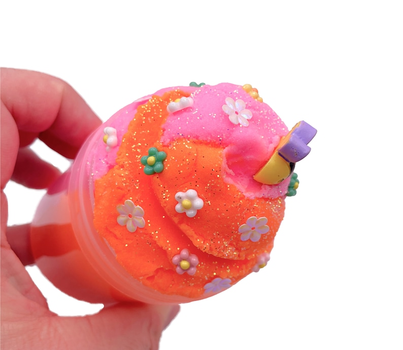 K&ouml;nnte beinhalten: Orangefarbener und rosafarbener Schleim mit Glitzer und wei&szlig;en Blumen in einem durchsichtigen Plastikbeh&auml;lter. Der Schleim ist mit einem gelben und violetten Blumenanh&auml;nger verziert.