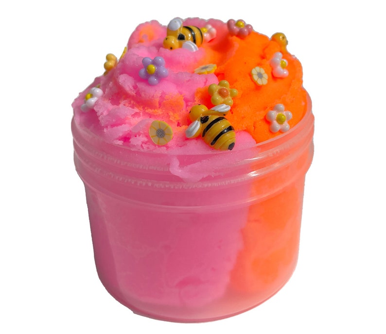 K&ouml;nnte beinhalten: Ein durchsichtiger Plastikbeh&auml;lter gef&uuml;llt mit zweifarbigem Schleim, unten rosa und oben orange. Der Schleim ist mit kleinen Plastikblumen und Bienen verziert.