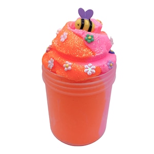 K&ouml;nnte beinhalten: Orangefarbener und rosafarbener Schleim in einem durchsichtigen Glas mit einer Biene und Blumenverzierungen oben drauf.