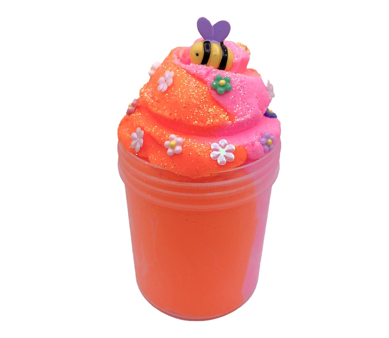 K&ouml;nnte beinhalten: Orangefarbener und rosafarbener Schleim in einem durchsichtigen Glas mit einer Biene und Blumenverzierungen oben drauf.
