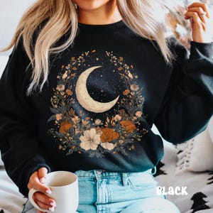 Op de afbeelding: Een zwart sweatshirt met een halve maan en een bloemenontwerp. De maan is omringd door verschillende bloemen en bladeren in tinten oranje, roze en wit. De tekst "Black" is zichtbaar in de rechterbenedenhoek van de afbeelding.