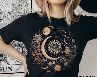 May include: Black t-shirt featuring a celestial design with a crescent moon, sun with a face, and floral elements. The design includes shades of beige, orange, and green. The word "BLACK" is printed in white at the bottom right.