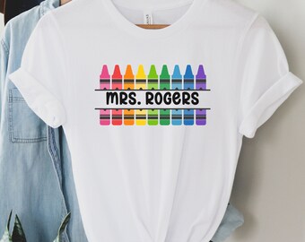 May include: White t-shirt with a rainbow row of crayons and the text "MRS. ROGERS" in black.