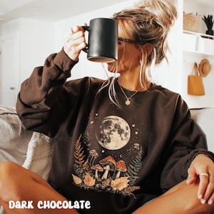 May include: A dark chocolate brown sweatshirt with a graphic print of a full moon, mushrooms, and leaves. The text "DARK CHOCOLATE" is printed on the bottom of the sweatshirt.