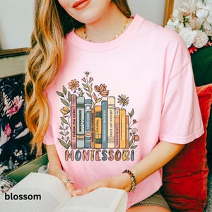 May include: Pink t-shirt with a colorful graphic of books and flowers. The word "MONTESSORI" is printed in a multi-colored font below the books. The shirt also features the word "blossom" at the bottom.