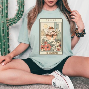 May include: A light blue t-shirt with a graphic of a cartoon ice cream cone with a cherry on top. The ice cream cone is wearing a dress and standing in a desert landscape with cacti. The text "The Icecream" is printed below the graphic.