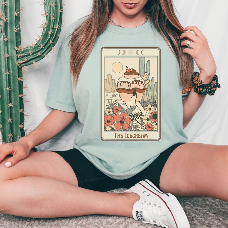 May include: A light blue t-shirt with a graphic of a cartoon ice cream cone with a cherry on top. The ice cream cone is wearing a dress and standing in a desert landscape with cacti. The text "The Icecream" is printed below the graphic.