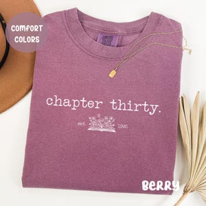 May include: A purple t-shirt with the text "chapter thirty. est. 1995" and a small illustration of a book with flowers on top. The shirt is made by Comfort Colors.