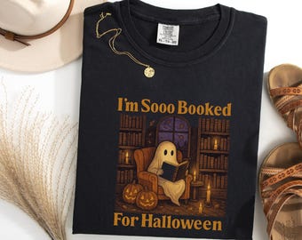 May include: Black crew neck t-shirt with the text "I'm Sooo Booked For Halloween". The graphic features a ghost reading in a library with pumpkins and candles. A gold necklace and a hat are next to the shirt.