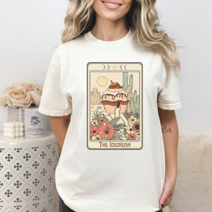 May include: A white t-shirt with a graphic print of a tarot card featuring an ice cream cone with a cherry on top. The card is titled "The Icecream".