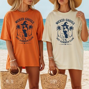 Op de afbeelding: Twee oversized T-shirts, één oranje en één crème, met de tekst "Wicked Ghouls Hot Summer Club" en een heksengrafiek. Het ontwerp bevat palmbomen en decoratieve elementen. De shirts worden gedragen met strooien draagtassen.