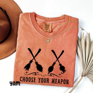 May include: An orange Comfort Colors t-shirt with a black graphic of two crossed witch brooms and the text "CHOOSE YOUR WEAPON". A gold necklace and a brown hat are also visible.