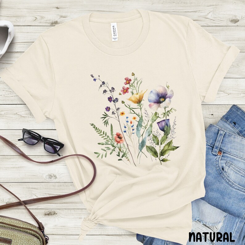 Op de afbeelding: Een beige T-shirt met een aquarel bloemenprint met paarse, blauwe, gele, rode en groene bloemen. Het shirt heeft een ronde hals en korte mouwen. Het woord "NATUURLIJK" staat in het zwart onderaan de afbeelding.