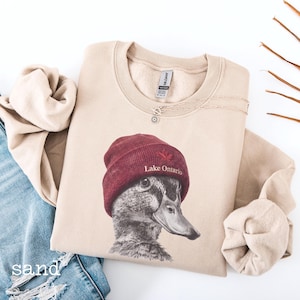 Op de afbeelding: Een zandkleurige sweater met ronde hals en een grafische print van een eend die een bordeauxrode muts draagt met een rood esdoornblad-icoon en de tekst Lake Ontario. De sweater heeft een geribbelde kraag en lange mouwen met boorden.