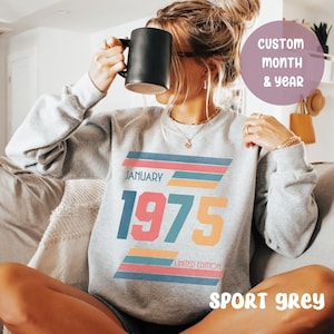 Op de afbeelding: Een grijs sweatshirt met een retro-ontwerp met de maand "januari" en het jaar "1975" in kleurrijke, gedurfde letters. Het ontwerp bevat kleurrijke strepen en de tekst "Limited Edition". Het sweatshirt wordt getoond op een model dat het draagt, en de tekst "Sport Grey" is zichtbaar onderaan de afbeelding.