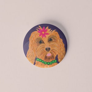 Puede incluir: Botón redondo con una ilustración de dibujos animados de un perro. El perro tiene pelo largo y lanudo de color marrón claro, una flor rosa y un collar de cuentas verdes. El fondo es azul oscuro.