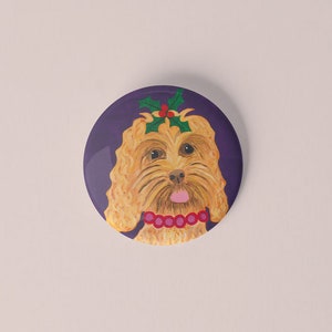 Puede incluir: Un botón redondo con una ilustración pintada de la cara de un perro. El perro tiene pelaje marrón claro, una lengua rosa y un collar rojo. Hojas y bayas de acebo adornan su cabeza, sobre un fondo morado.