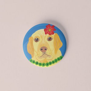 Può includere: Un bottone rotondo dipinto raffigurante un Labrador Retriever giallo con occhi marroni, un fiore rosso e una collana di perline verdi. Il cane è ambientato su uno sfondo blu.