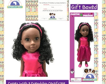 Puede incluir: Una muñeca rosa y negra con cabello negro rizado que lleva un vestido rosa con una flor en el frente. La muñeca viene con un certificado de amistad y está en caja de regalo. La caja tiene las palabras "Positive & Precious" en ella.