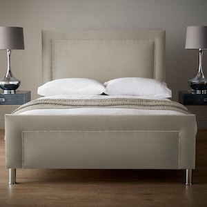 May include: A beige upholstered bed with a matching headboard and silver stud detailing. Two bedside tables with silver lamps flank the bed. The bed is made up with white bedding and a beige throw. The bed frame is supported by silver legs.
