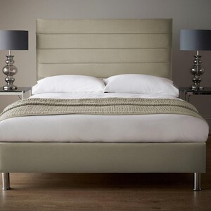 May include: A light grey upholstered bed frame with a tall headboard featuring horizontal channel tufting. The bed is made with white linens and a grey throw blanket. Two bedside tables with glass tops and grey lampshades are on either side of the bed.