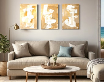 May include: Three abstract paintings in a set, each with a white geometric design on a beige background. The paintings are hung on a wall above a beige sofa and a round coffee table.