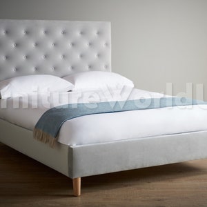 May include: A grey upholstered bed frame with a tufted headboard and wooden legs. The bed is made with white bedding and a blue throw blanket.