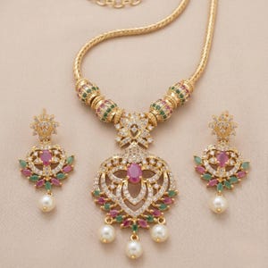 Puede incluir: Un conjunto de joyas dorado que incluye un collar y pendientes a juego. El collar tiene un colgante con una piedra rosa central, rodeada de cristales transparentes y piedras rosas y verdes más pequeñas. Los pendientes reflejan el diseño del colgante, con una perla colgante.