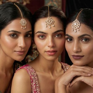 May include: Three women wearing gold-toned headpieces with intricate designs and dangling accents. The headpieces are centered on the forehead, with delicate chains extending to the hair. The women are wearing pink and gold embellished tops.