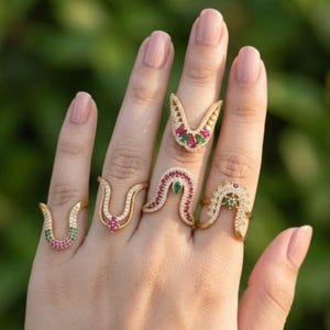 May include: A hand wearing five gold-toned rings. Each ring features a unique design with colorful gemstones, including red, green, and clear stones. The rings are set against a blurred green background.