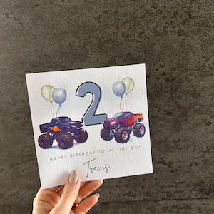 May include: A birthday card with two monster trucks, a number 2, and balloons. The card reads "HAPPY BIRTHDAY TO MY TINY GUY" and includes the name "Travis". The trucks are purple and red.