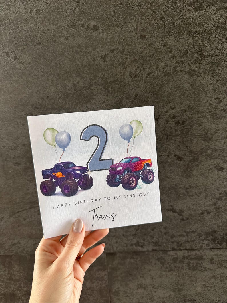 May include: A birthday card with two monster trucks, a number 2, and balloons. The card reads "HAPPY BIRTHDAY TO MY TINY GUY" and includes the name "Travis". The trucks are purple and red.