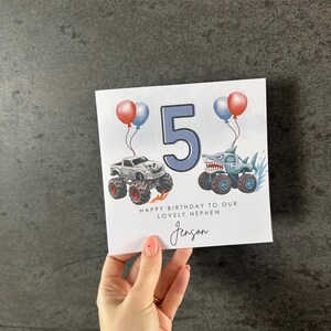May include: A birthday card featuring the number 5 in blue, two monster trucks, and balloons. The card says "Happy Birthday to our lovely nephew Jenson."