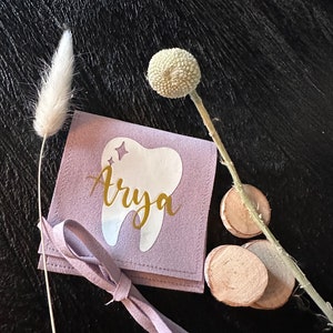 May include: A lavender-coloured pouch with a white tooth graphic and the name "Arya" written in gold lettering. The pouch has a tie closure and is sitting on a dark wooden surface.