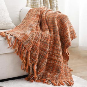 May include: A woven throw blanket in shades of orange and brown, with a fringed edge, draped over a white sofa. The blanket's texture is visible, with a mix of orange, brown, and white threads. The fringe is a mix of orange and cream.