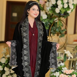 May include: A woman wearing a black velvet shawl with silver embroidery. The shawl is draped over a maroon dress. The woman is standing in front of a floral arrangement.