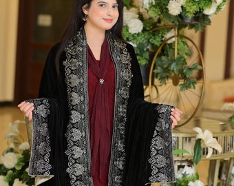 May include: A woman wearing a black velvet shawl with silver embroidery. The shawl is draped over a maroon dress. The woman is standing in front of a floral arrangement.