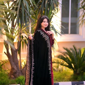 May include: A woman wearing a black velvet shawl with silver embroidery and a maroon dress. She is standing in front of a window and a palm tree.
