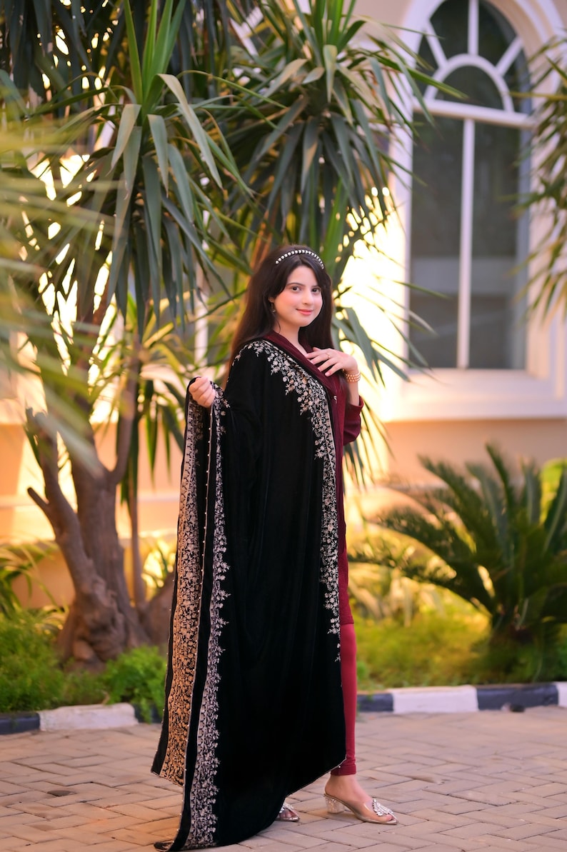 May include: A woman wearing a black velvet shawl with silver embroidery and a maroon dress. She is standing in front of a window and a palm tree.