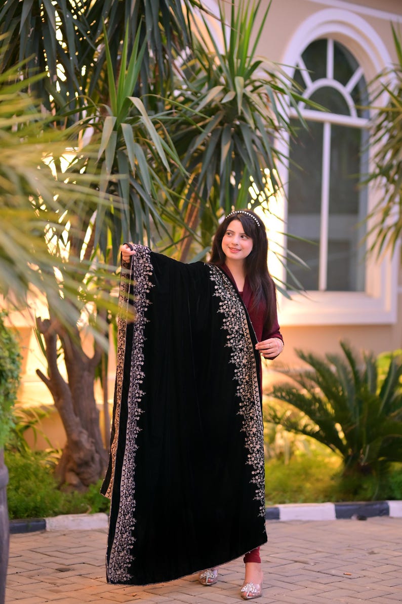 May include: A woman wearing a maroon dress and a black velvet shawl with gold embroidery. The shawl has a decorative border with intricate floral patterns. She is standing in front of a lush green plant and a building with a window.