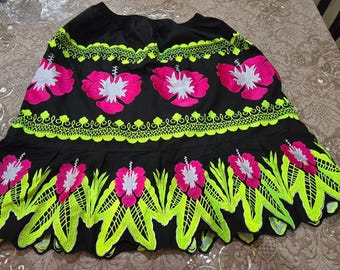 May include: Black skirt with vibrant neon green and pink floral embroidery. The skirt features large hibiscus flowers and decorative trim, creating a bold and colourful design. The skirt is made of a flowing fabric.