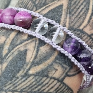 May include: A handmade bracelet featuring a macrame design in a light purple color. The bracelet is adorned with a combination of round, polished beads in shades of pink, clear, and deep purple. The bracelet is a piece of jewelry.