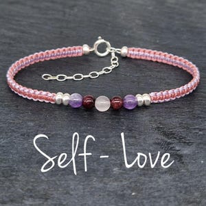 Puede incluir: Un brazalete trenzado de color rosa y morado con un cierre plateado y una fila de cuentas de amatista, granate, cuarzo rosa y granate. El brazalete tiene el texto "Self-Love" escrito debajo.