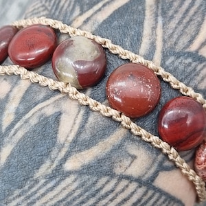 May include: A red and brown stone bracelet with a beige cord. The bracelet is made with round stones and has a braided cord.