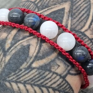 May include: A red braided bracelet with white and dark blue round beads. The bracelet is worn on a person's wrist.